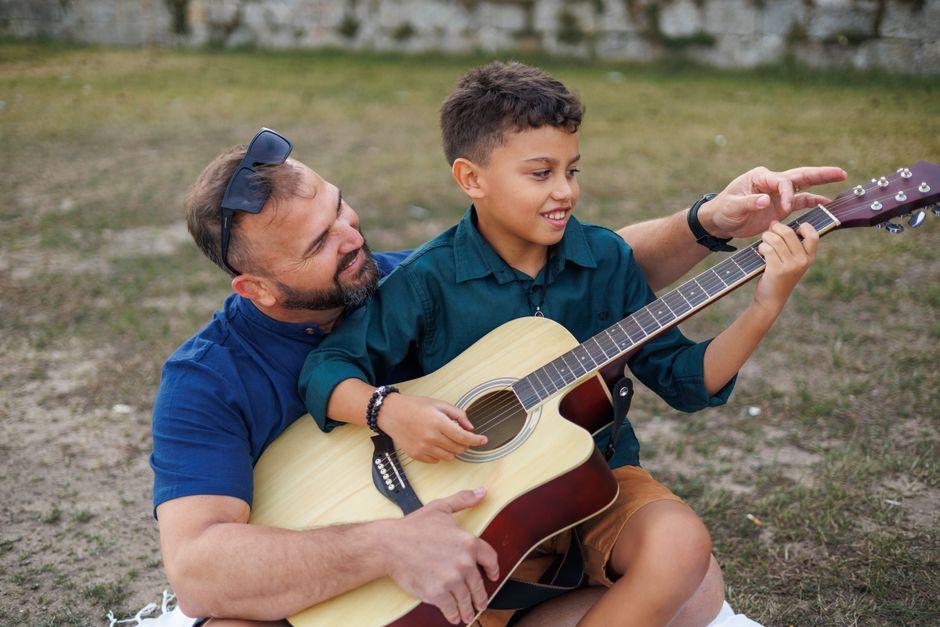 Comment trouver un bon professeur de guitare en Belgique : critères, tarifs et méthodes d'apprentissage 7 Professeur de guitare guidant les doigts d'un enfant sur le manche d'une guitare acoustique en extérieur